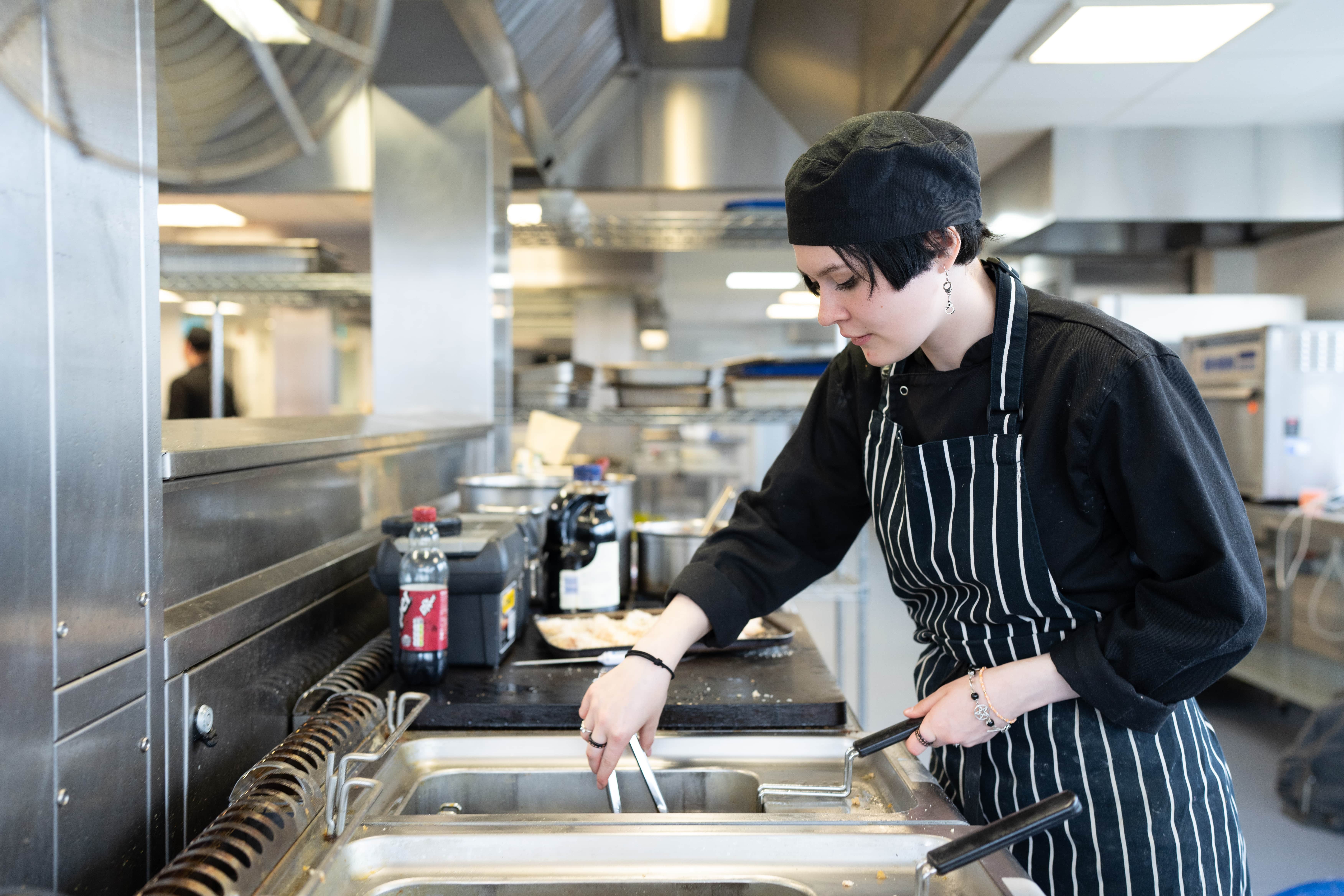 student uses tongs to pull something out of fryer in training kitchen