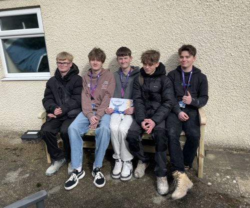 5 plastering students sat on the bench they made smiling, with 1 student holding a box of shortbread biscuits for the residents