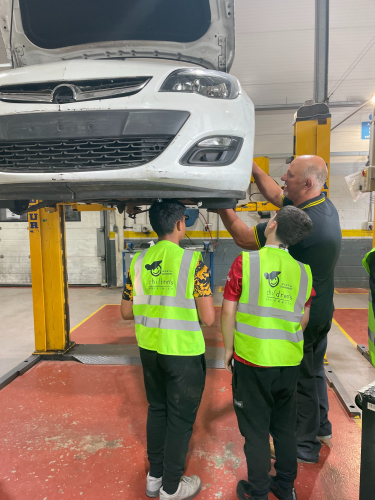 Two students being taught how to fix the wheel of a car by a member of staff